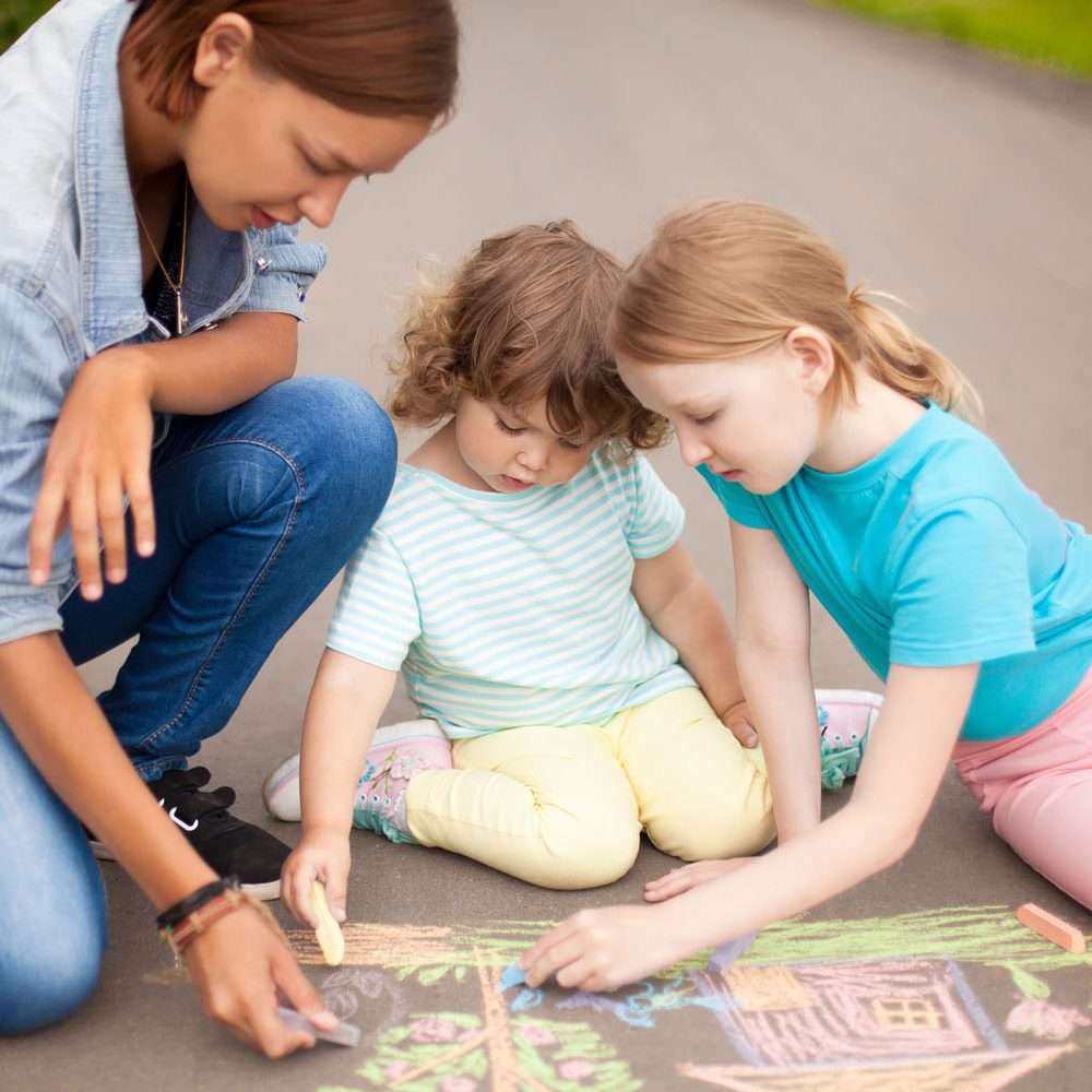 Little girls chalking at the asphalt with elder sister or nanny. Sisters friendship. Best friends children happy family. Drawing with color chalk. Happy childhood. Kids in kindergarten. Preschooler leisure time. Kids spending time without parents. Childre